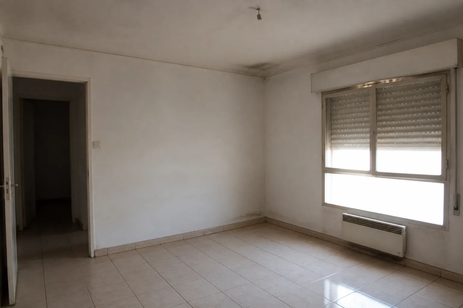 Pièce d'appartement inoccupé avec volets mi-clos et traces de moisissure dans un angle
