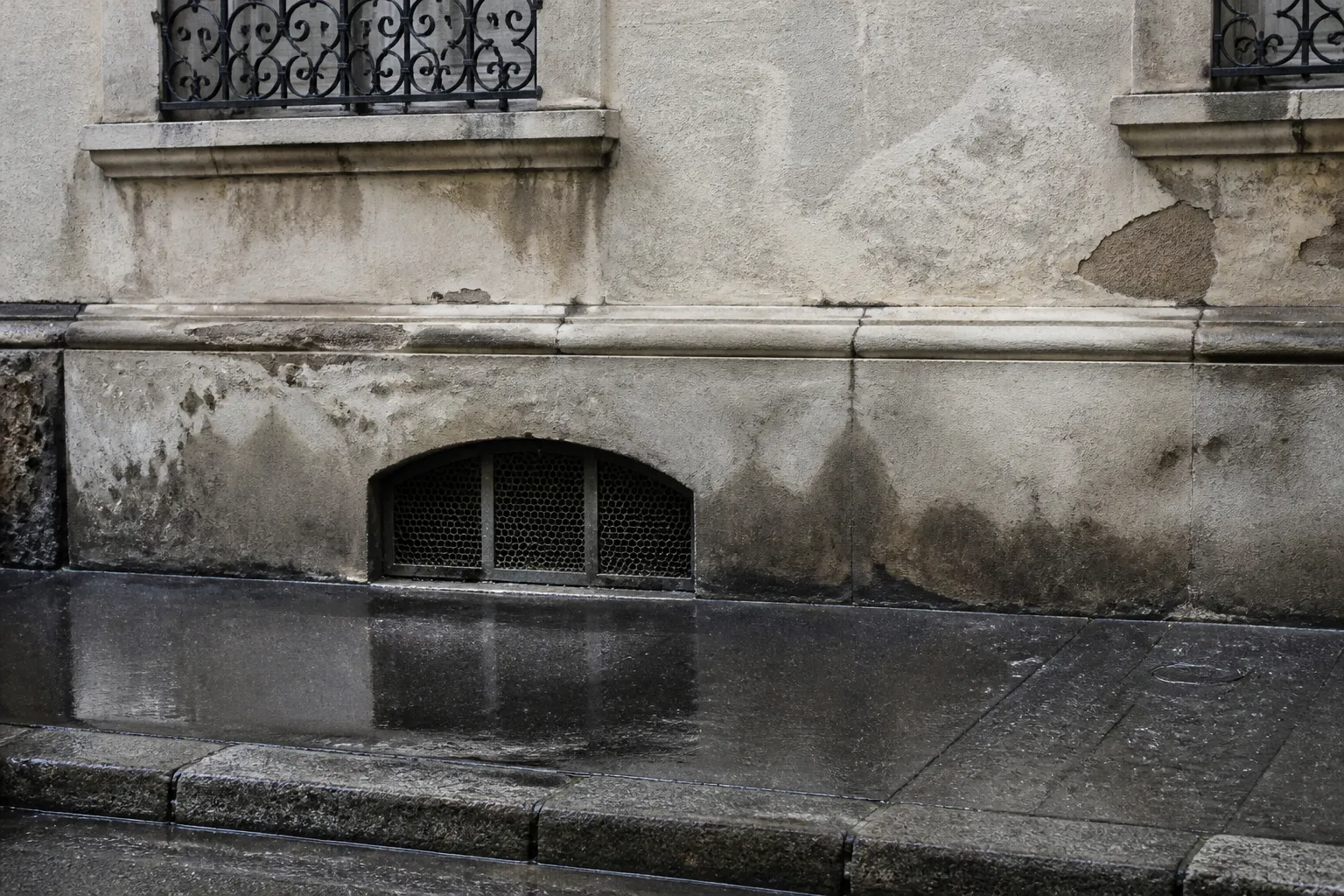 Pied de façade avec soupirail et enduit marqué par l’humidité après la pluie