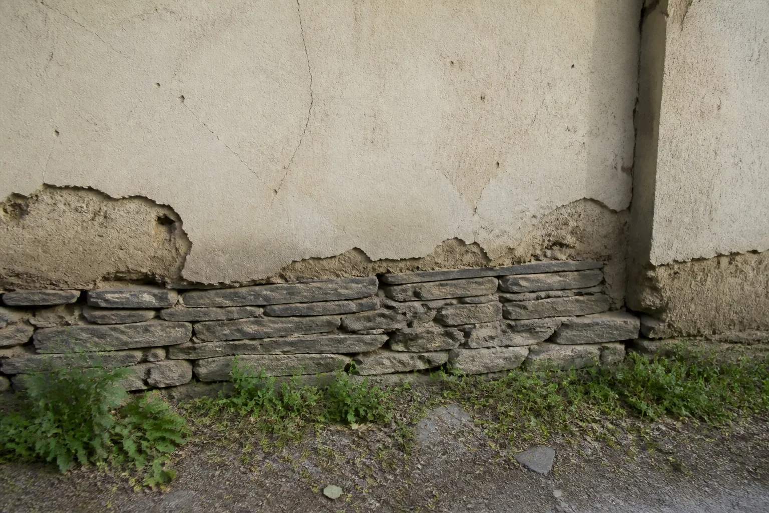 Mur ancien en terre crue sur soubassement de pierre, enduit à la chaux dégradé en pied de mur